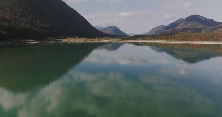 A calm lake reflects the surrounding mountains under a partly cloudy sky