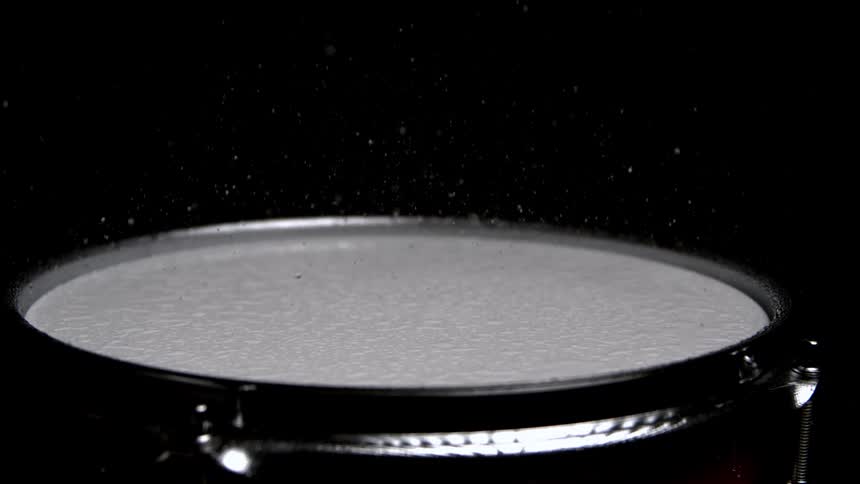 A wooden drumstick rests on a white drumhead as fine powder rains down sending a misty burst of particles into the air against a dark background