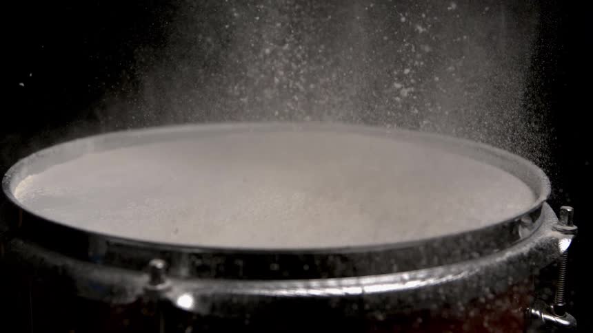 A drumcovered in a layer of fine white powder and a drumstick strikes the top illustrating the kinetic energy and subsequent vibrations in a powerful plume of dust against a stark black background