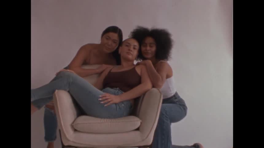 Three women pose closely together in a studio setting with one seated in a chair and the other two embracing her from behind they share a confident and affectionate moment while looking directly at the camera