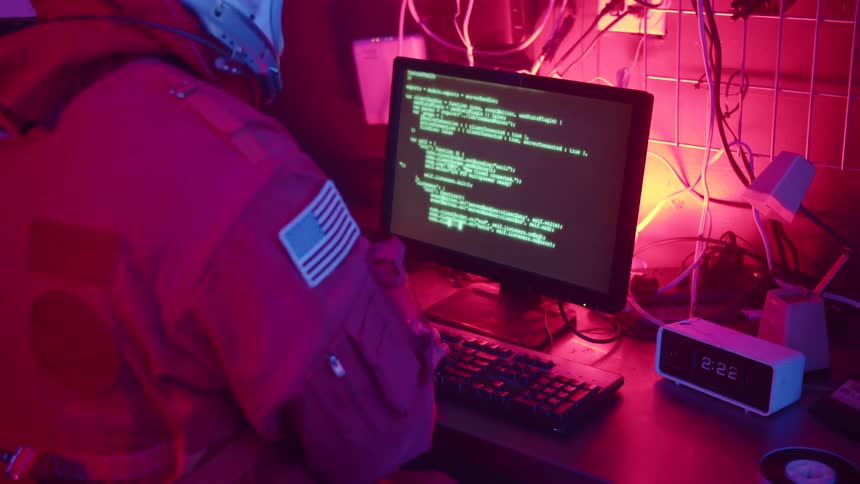 A person wearing a spacesuit types code on a computer inside a room illuminated by red light
