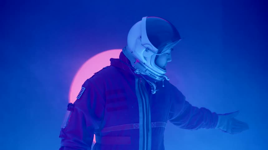 A young woman wearing a spacesuit and helmet stands in a dark room illuminated by blue light