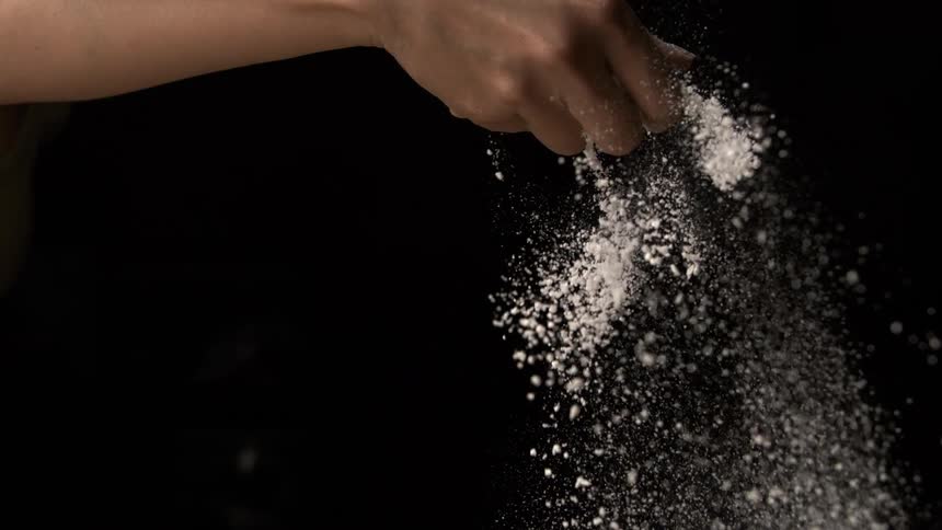 The video is a highcontrast closeup of a persons hand sifting or sprinkling a large amount of white powder likely flour or sugar against a pitch black backgroundthe powder falls in a bright cascade with individual granules clearly visible agains