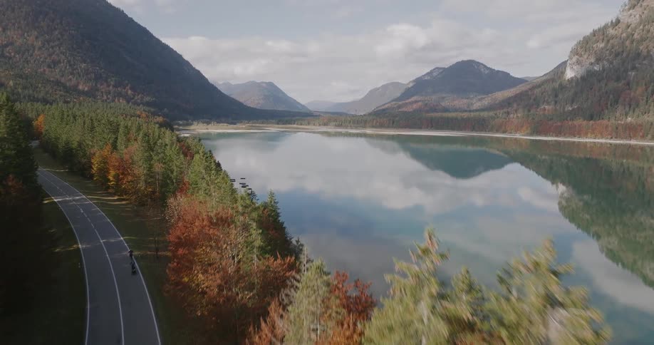 The video shows an elevated view of a narrow paved road winding through an autumn forest beside a calm lakethe camera rises revealing a stunning panorama of the serene lake reflecting the surrounding mountains and the colorful fall foliage