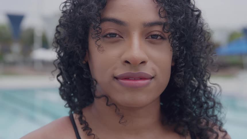 A young black woman with long curly hair standing in an outdoor pool area at midday