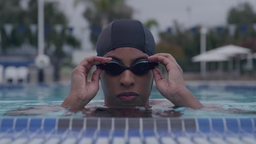 The video is a closeup shot of a female swimmer emerging from the water at the edge of a pool wearing a black swim cap and goggles she adjusts her goggles with a focused and determined expression suggesting she is preparing for a race or intense train