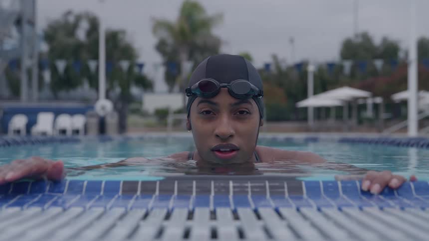 Androgynous person wearing a swim cap and goggles staying on the edge of a swimming pool outdoors during day time