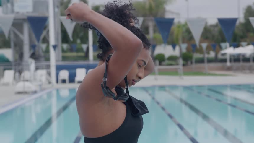 The video shows a swimmer standing on the edge of an outdoor pool as she secures her goggles over her head in preparation for swimming she is wearing a dark swimsuit and the pools lanes and surrounding flags are visible in the bright background