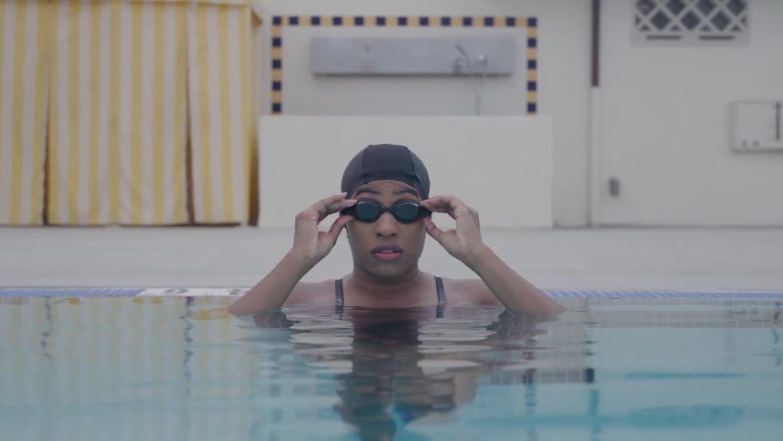 A swimmer in a pool adjusts her goggles while looking directly at the camera with a striped curtain and poolside equipment in the background
