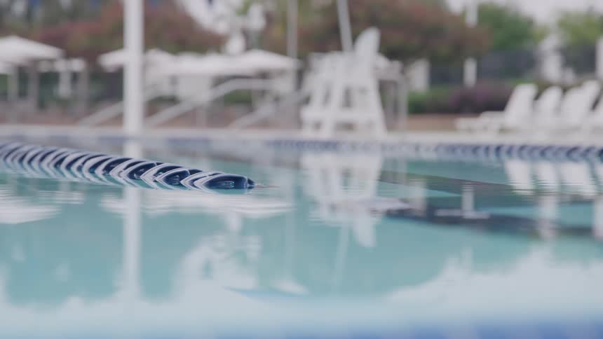 A dark blue lane line floats on the surface of an empty outdoor swimming pool where the clear water gently reflects the cloudy sky in the blurred background white umbrellas and deck chairs surround the edge suggesting a quiet recreational area