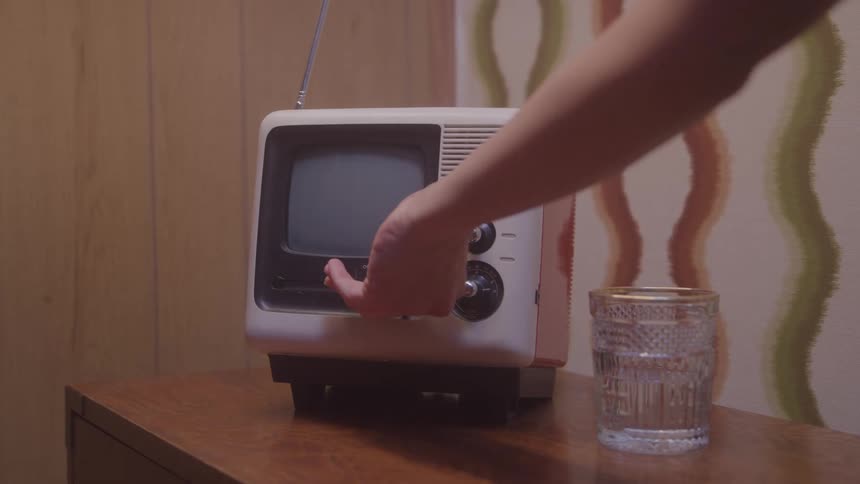A person reaches to turn on a small vintage television on a wooden table