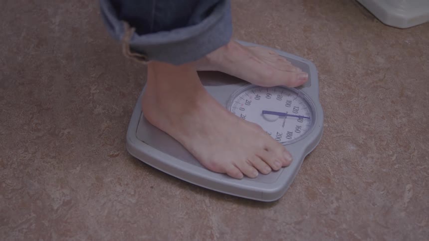 The video is a closeup of a persons bare feet stepping onto a standard analog bathroom scalethe dial immediately moves indicating that the person is weighing themselves while wearing rolledup blue jeans