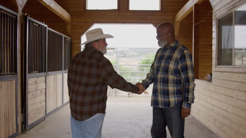 Two older men one wearing a cowboy hat and a brown plaid shirt and the other wearing a blue plaid shirt face each other and exchange a firm handshake inside the wooden hallway of a horse stable with sunny green hills visible in the background