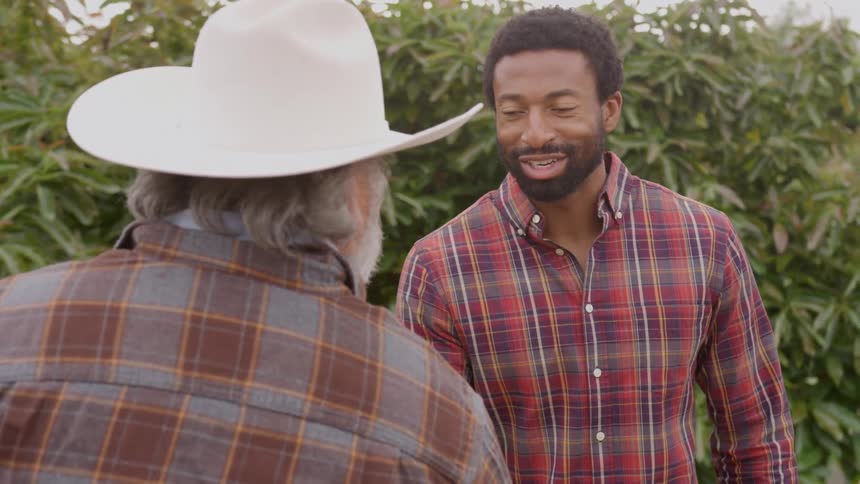 Two men engaged in a friendly conversation in an outdoor setting filled with green foliage as the younger man wearing a red plaid shirt smiles genuinely while looking at the older man who is seen from the back wearing a white hat and a plaid shirt