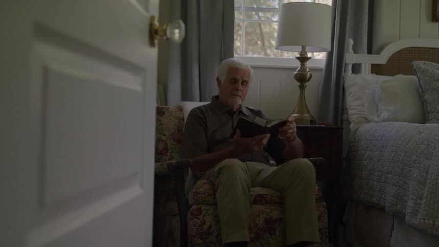 An elderly man with white hair and a beard is sitting in a floral armchair focused on a small electronic device he is positioned in a cozy bedroom with a window and a made bed visible behind him