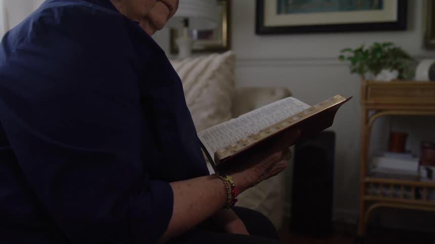 An elderly woman sitting on her couch reading the bible in her hands at midday