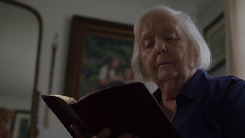 The video features a closeup on an elderly woman with short white hair and a blue shirt intently reading a book with a dark red or maroon covershe is framed against a softfocus background of what appears to be a living room with a mirror and frame