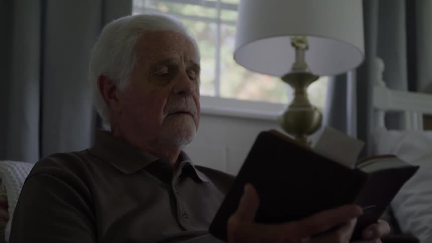 An elderly man reads a book by the window in a softly lit room