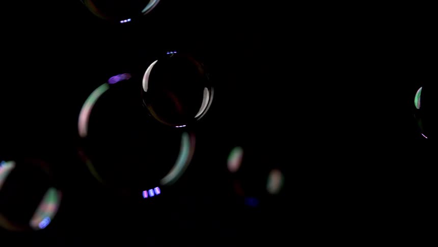 The video shows several soap bubbles floating slowly across a completely dark black background the bubbles reflect rainbowcolored spectral highlights from an unseen light source giving them an iridescent and ethereal glow as they drift