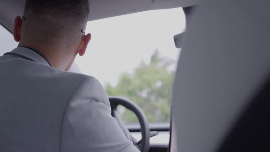 A man in a light gray suit drives in a vehicle while glancing to the left