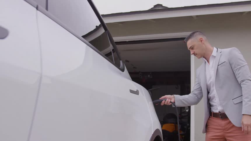 A man in business attire charges a white electric car outside a garage