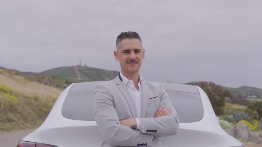 Man stands behind his parked car with his arms folded as he turns his head