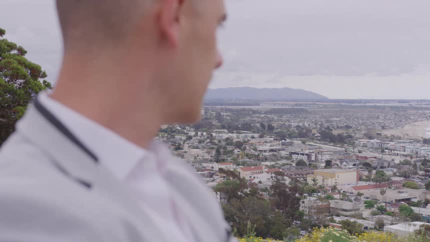 A man standing on a hill watches the city skyline in the background at midday
