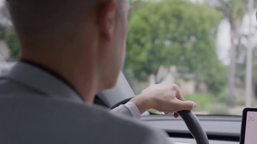 Man holds one hand on the steering wheel as he drives through a neighborhood