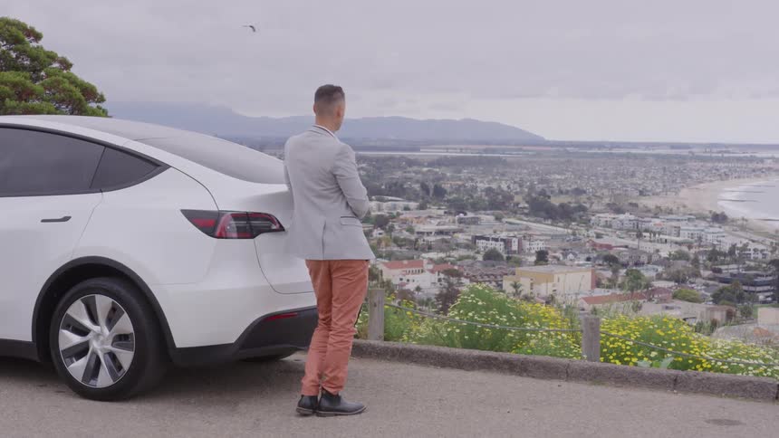 A man in a light jacket and orange pants stands next to the back of a white suv overlooking a sweeping coastal city vista the viewpoint is high above the town with the ocean beach and distant mountains visible beneath an overcast sky