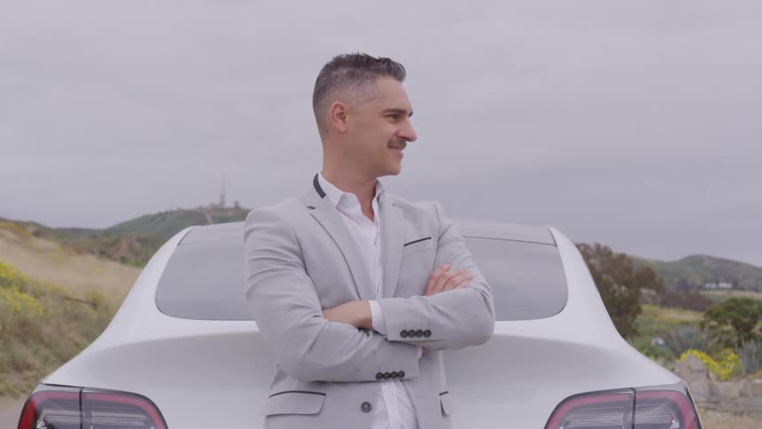A smiling man with slickedback hair stands with his arms crossed leaning against the back of a white vehicle he is wearing a light gray jacket and is set against a muted landscape with a cloudy sky