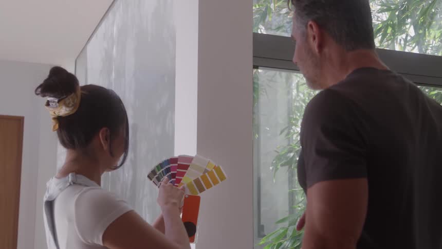 A woman and a man are standing in a bright room looking at a wall near a large windowthe woman holds up a fanshaped paint swatch deck comparing the colors against a light gray wall next to a white wall