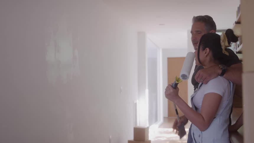 A couple stands close together in a hallway looking at a wall with fresh white paintthe man has his arm around the woman and points toward the wall appearing to discuss their plans for the new room