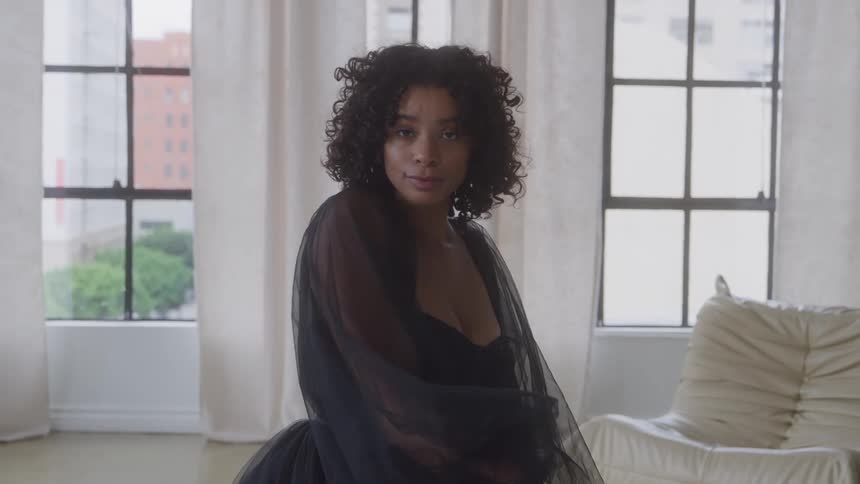 Curlyhaired woman wearing a sheer black outfit poses in a room lit by large windows