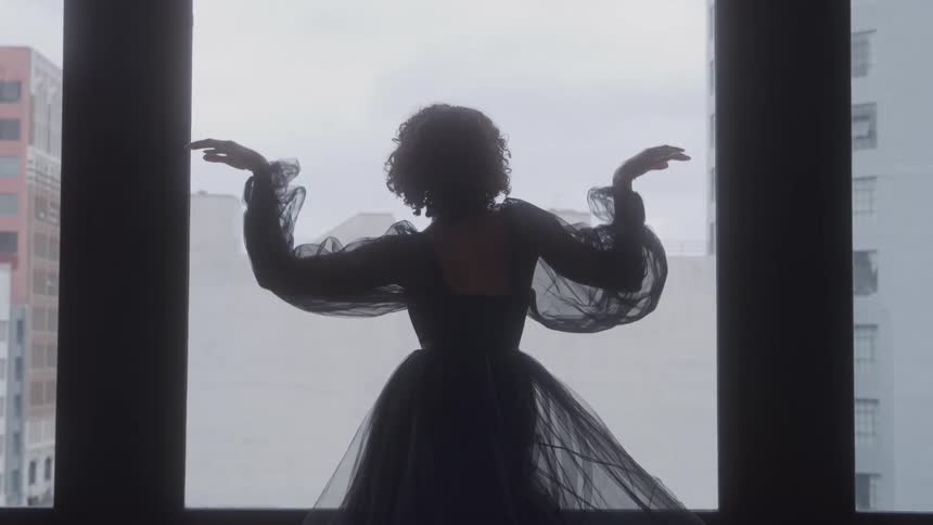 Woman in a sheer dress raises her arms in front of a bright window