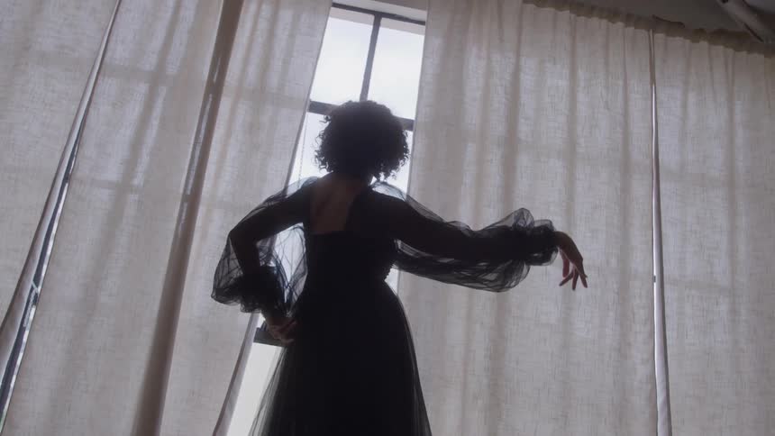 The video shows a woman in silhouette with curly hair wearing a long dark flowing dress with sheer puffed sleevesshe is dramatically posed in front of a bright window covered by sheer lightcolored curtains with her back mostly to the camera and