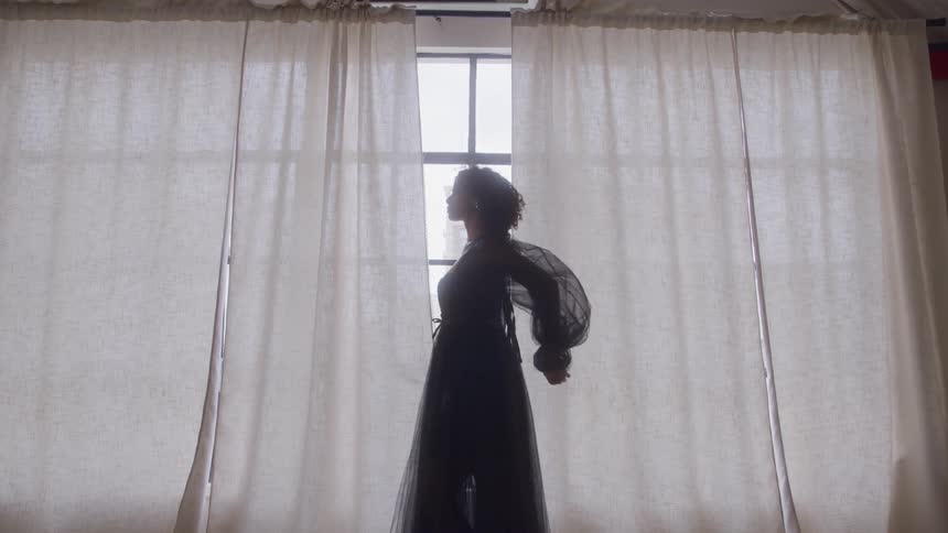 A woman is dancing near a large window with curtains