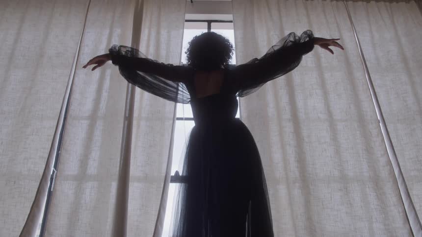 A woman standing at a large window slowly raises her arms outward