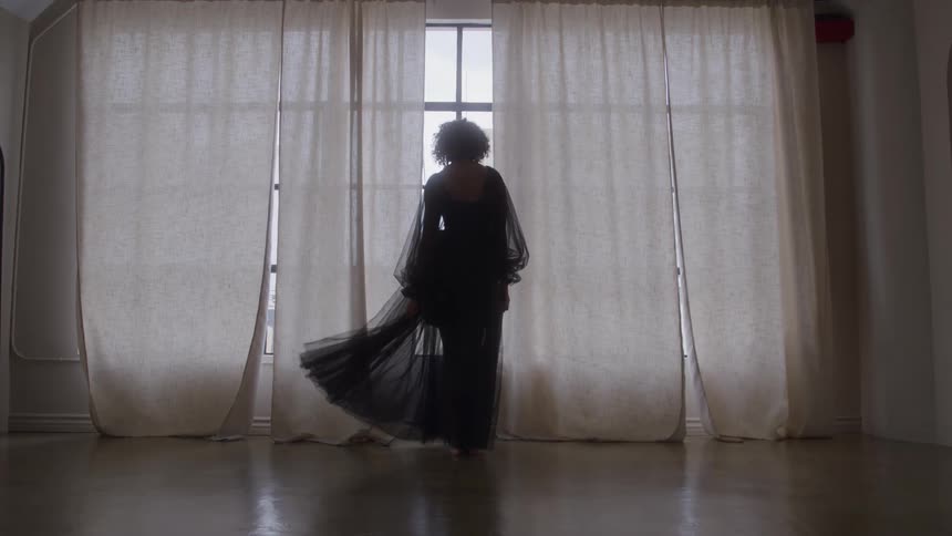 The video shows a dramatic shot of a woman silhouetted against a large window framed by two long sheer white curtains she is wearing a flowing darkcolored gown and briefly turns or sways allowing the fabric to billow out around her suggesting a dan