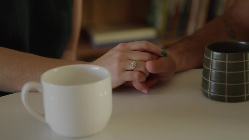 A couples hands clasp gently beside a white mug and a patterned dark mug on a table