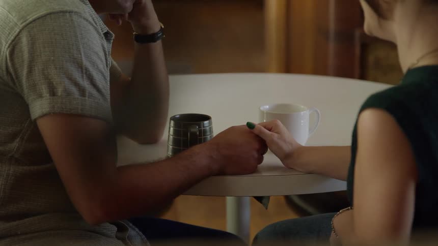 A man and a woman sit at a small round table holding hands beside two coffee mugs sharing a quiet supportive moment