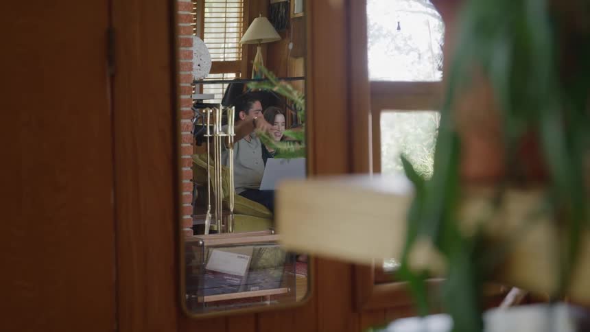 Mirror reflects two people sitting on a couch with a laptop smiling and talking