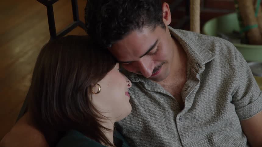 A young couple sitting on the living room couch smiling and leaning their heads against each other at midday