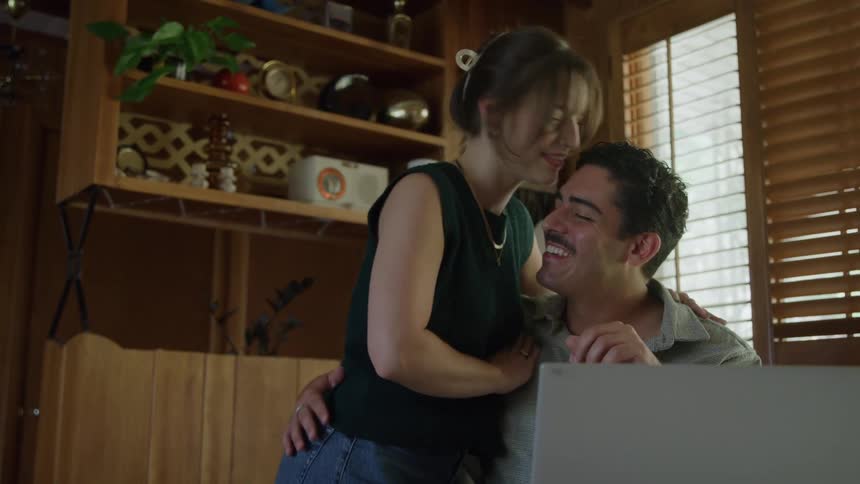 A woman rests her hands on a mans shoulders as he smiles at a laptop in a cozy sunlit room
