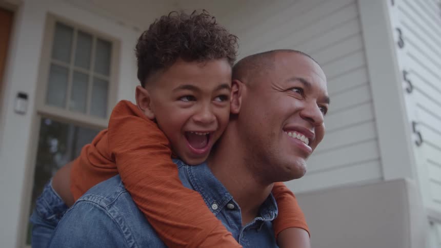 Father carries his son on his back as they both smile in front of the house