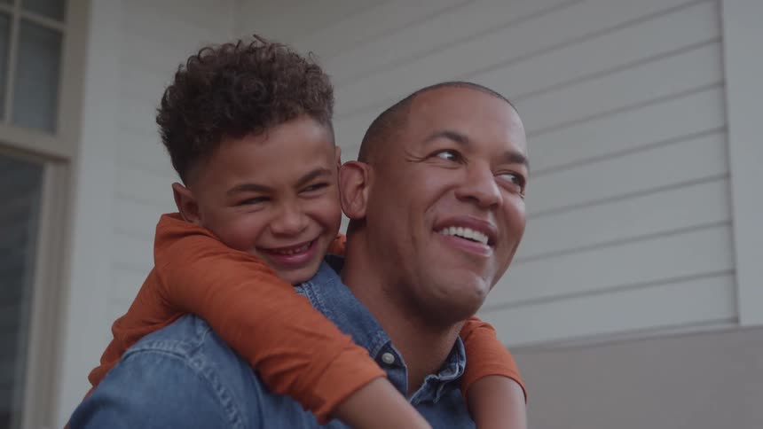 This is a heartwarming closeup shot of a father and son together on a porch or deckan adult man in a denim shirt is giving a piggyback ride to a young boy in an orange shirtboth are smiling widely and laughing conveying a feeling of joy and loving