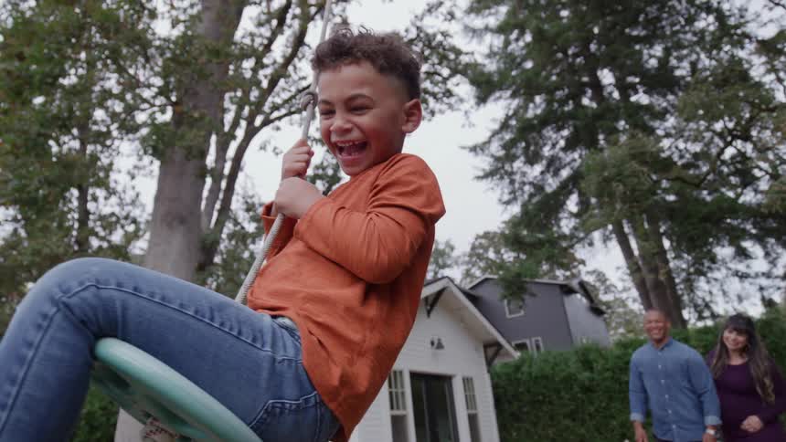 A boy swings on a rope while his parents smile nearby in their backyard
