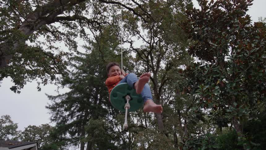 A child on a rope swing soars barefoot facing forward against a backdrop of tall trees