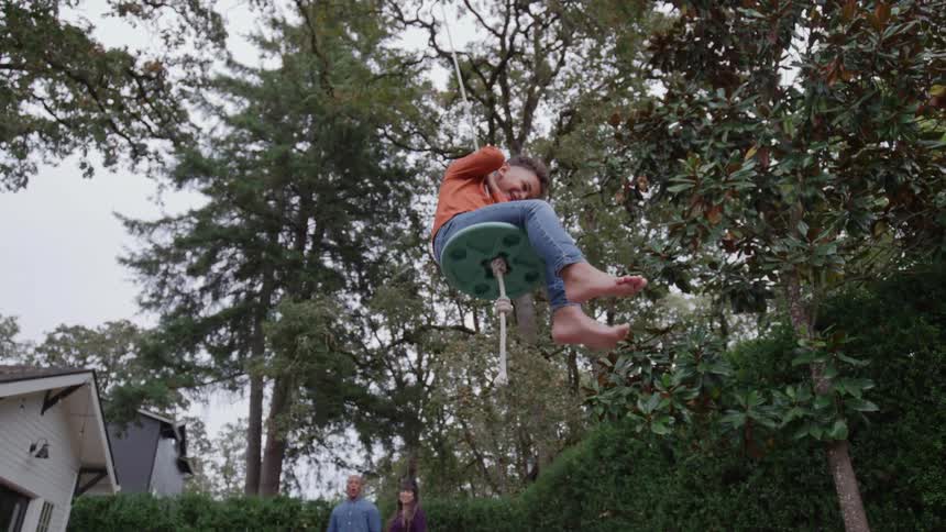 A child swings high on a rope swing while parents watch behind in a lush backyard