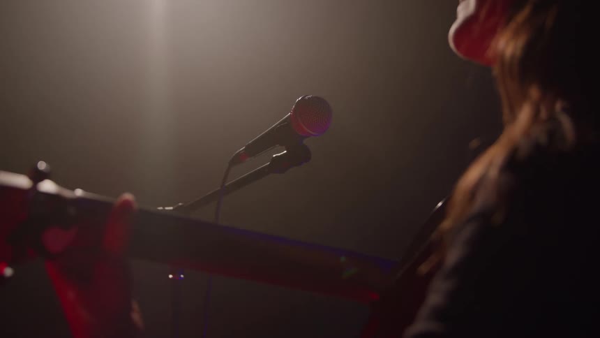 A person is shown from a low angle on stage playing an acoustic guitar into a microphone stand the scene is darkly lit and dramatic with a beam of light hitting the performance area from above