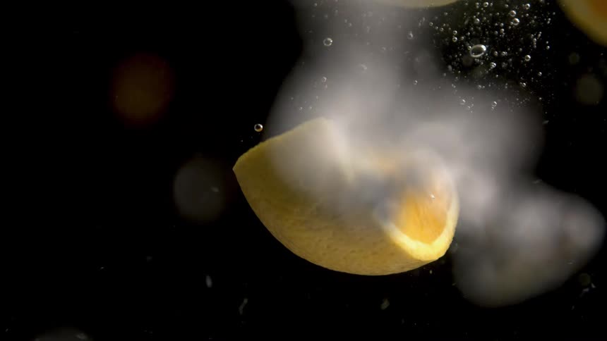 A vibrant yellow citrus fruit wedge is captured in extreme slow motion as it descends through a dark effervescent liquid generating a stream of small distinct bubbles as it settles slowly into the frame
